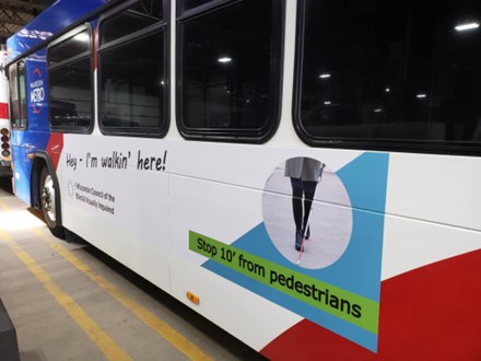 An ad on the side of a Waukesha bus featuring a photo of the lower half of a person walking with a white cane, the Council logo, and the words Hey, I'm walkin' here and Stop 10 feet from pedestrians.