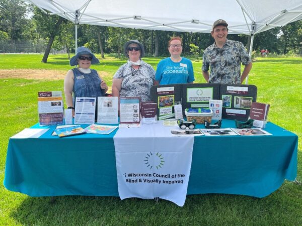 Four smiling people standing outdoors behind a table bearing Wisconsin Council of the Blind & Visually Impaired information.