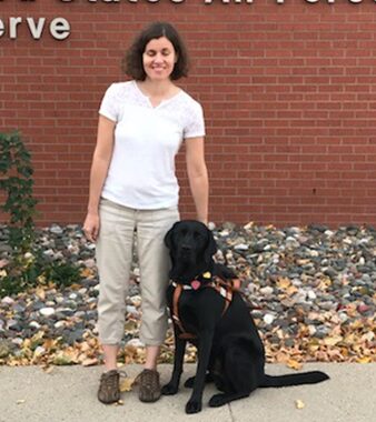 A light-skinned woman standing next to a sitting guide dog