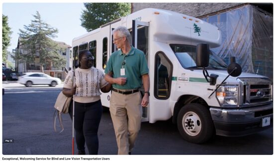 A light-skinned man using human guiding technique to assist a dark-skinned woman holding a white cane as they walk away from a paratransit vehicle.
