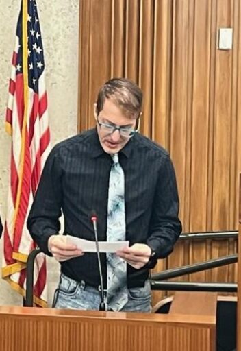 A light-skinned man speaking at a podium with an American flag behind him