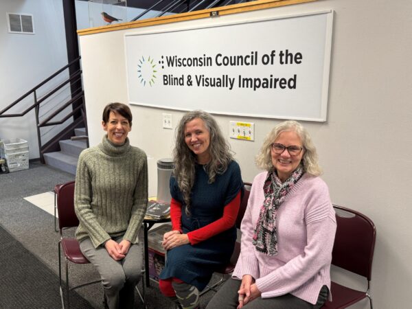 Three light-skinned women sitting in chairs in front of a sign reading Wisconsin Council of the Blind & Visually Impaired