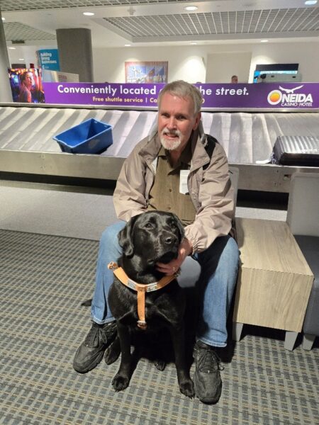 A light-skinned man sitting on a bench with a guide dog in front of a baggage claim carousel.