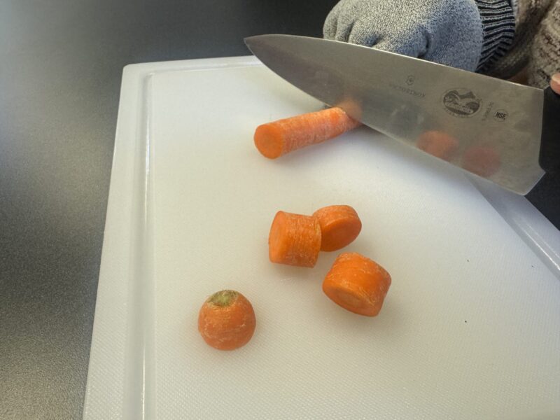 Two hands chopping a carrot with a knife on a cutting board.
