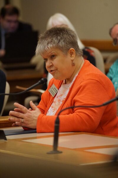 A light-skinned woman sitting at a table speaking into a microphone.