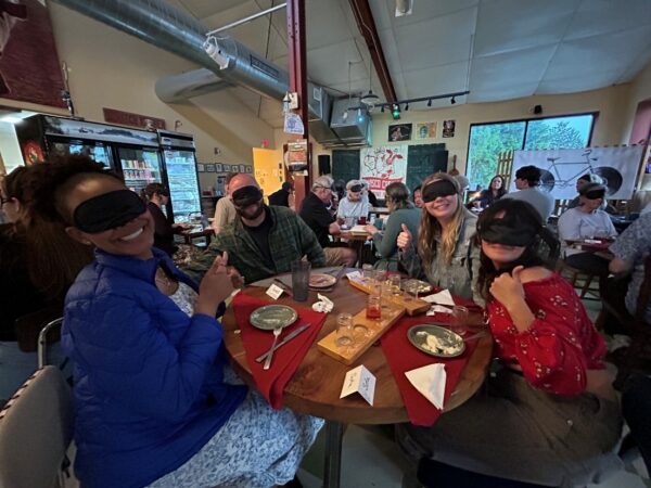 Four blindfolded people sitting at a round table with finished plates of food in front of them smiling toward the camera