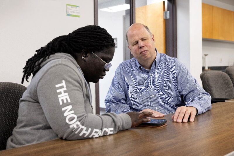 A dark-skinned woman using a smart phone as a light-skinned man observes.