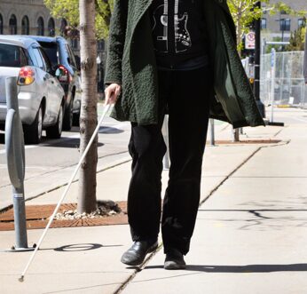 The lower half of a person using a white cane to navigate a city sidewalk.