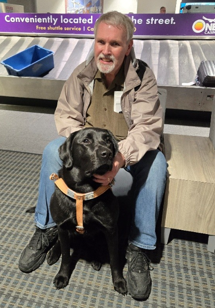 A light-skinned man sitting on a bench with a guide dog in front of a baggage claim carousel.