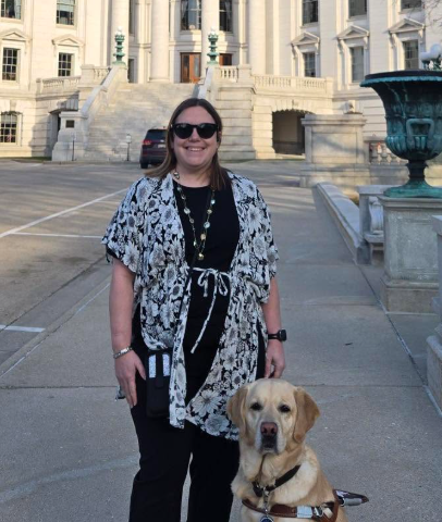 A light-skinned woman standing with her guide dog in front of the Wisconsin State Capitol steps