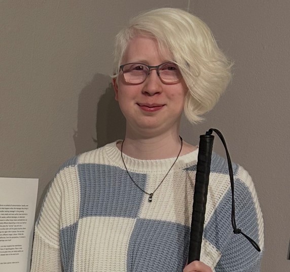 A light-skinned person holding a cane standing against a wall