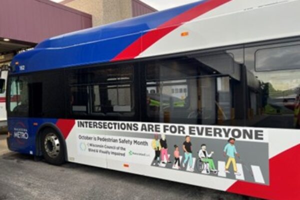 A Waukesha Metro bus with a large ad on the side reading Intersections Are For Everyone, October is Pedestrian Safety Month with a graphic showing a diverse group of people crossing at an intersection
