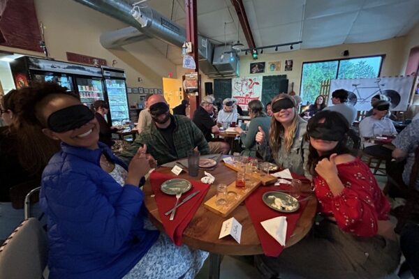 Four blindfolded people sitting at a round table with finished plates of food in front of them smiling toward the camera